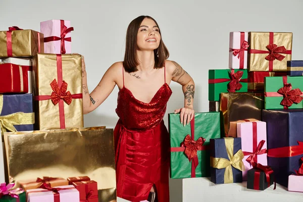 Jeune femme souriante en tenue de fête debout à côté de cadeaux regardant loin, concept de cadeaux de vacances — Photo de stock