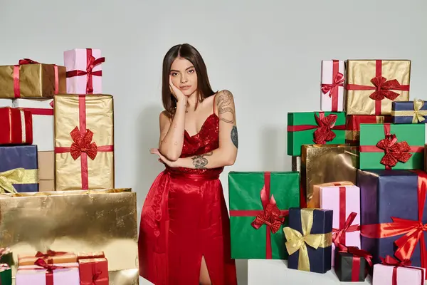 Femme rêveuse en robe rouge de fête posant à côté de cadeaux avec la main sur la joue, concept de cadeaux de vacances — Photo de stock