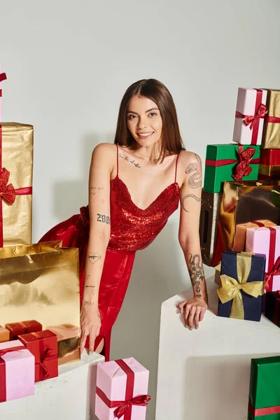 Attrayant femme en robe de fête avec des tatouages posant avec pile de cadeaux, concept de cadeaux de vacances — Photo de stock