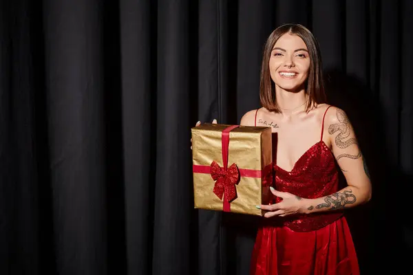 Fröhliche Dame lächelt in die Kamera mit Geschenk in der Hand mit schwarzen Vorhängen vor dem Hintergrund, Weihnachtsgeschenke — Stockfoto