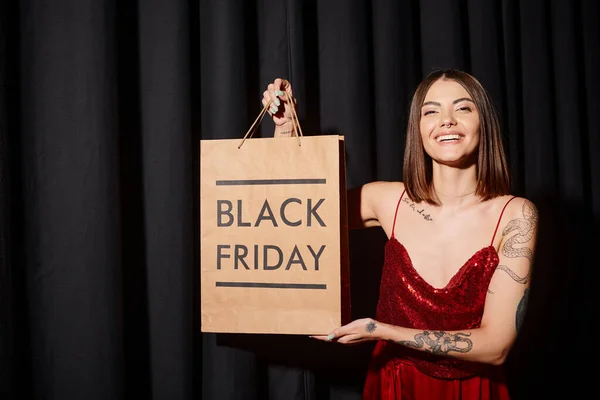 Pretty woman with shopping bag smiling at camera with black curtains on backdrop, black friday — Stock Photo