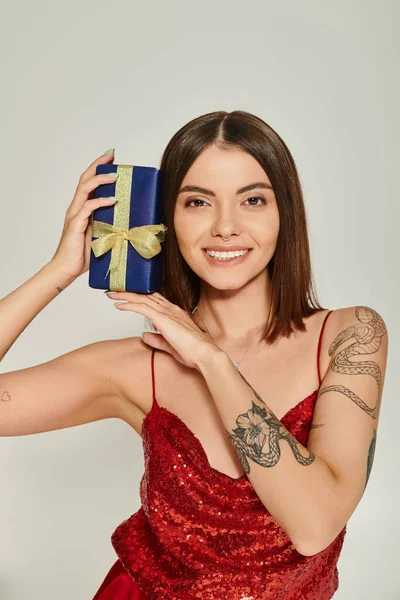 Cheerful lady with tattoos and piercing holding present near face smiling at camera, holiday gifts — Stock Photo