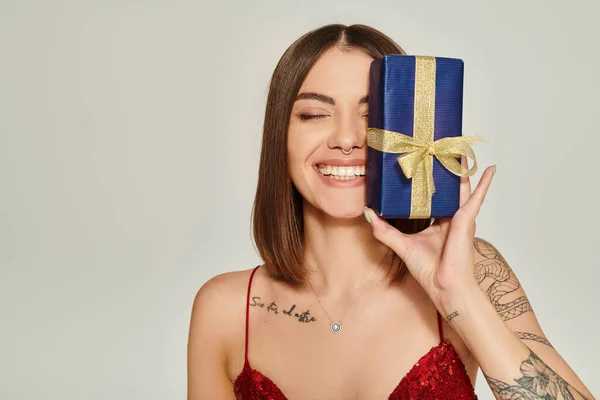 Mulher atraente segurando presente perto de seu rosto e sorrindo sinceramente com olhos fechados, presentes de férias — Fotografia de Stock