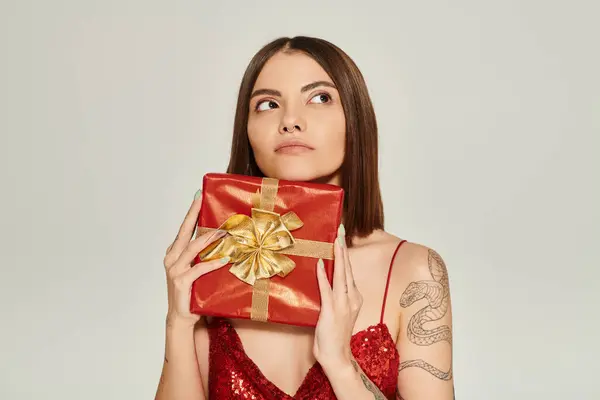 Mujer joven reflexiva sosteniendo presente rojo y soñando mirando hacia otro lado, concepto de regalos de vacaciones - foto de stock