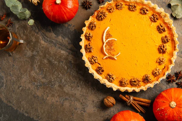 Thanksgiving still life, garnished pumpkin pie near ripe pumpkins, herbs and tea on stone surface — Stock Photo