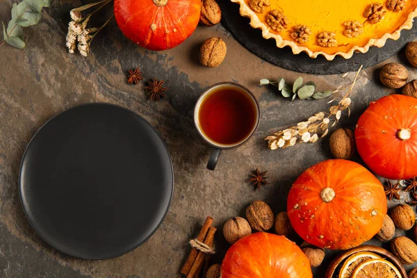 Piatto in ceramica nera vicino alle zucche arancioni, tè e torta di zucca del Ringraziamento su un tavolo in pietra rustica — Foto stock