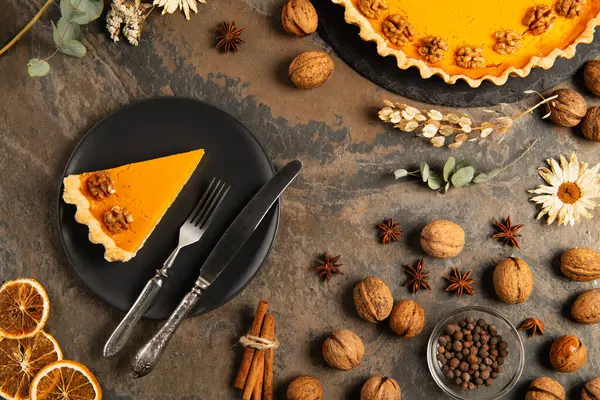 Вкусный тыквенный пирог и столовые приборы на черной керамической тарелке рядом с апельсиновыми тыквами на каменном столе, День благодарения — стоковое фото