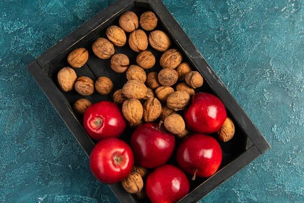 Red ripe apples and whole walnuts in black wooden tray on blue textured table, thanksgiving concept — Stock Photo