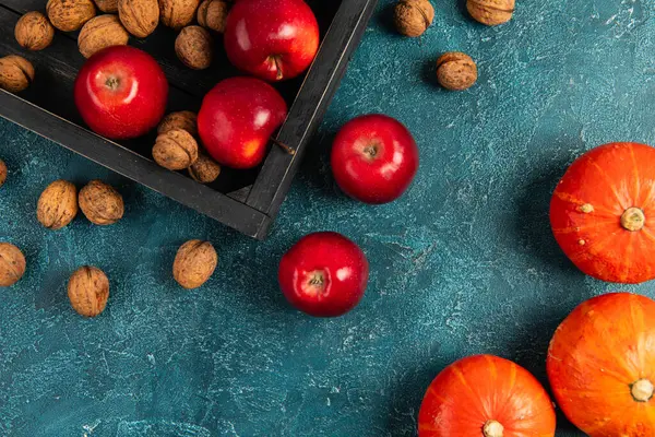 Помаранчеві гарбузи біля чорного лотка з волоськими горіхами та червоними яблуками на синьому текстурованому фоні, подяка — стокове фото