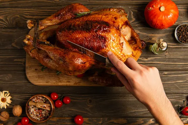 Cropped view of man cutting thanksgiving turkey near cherry tomatoes and pumpkin on wooden table — Stock Photo