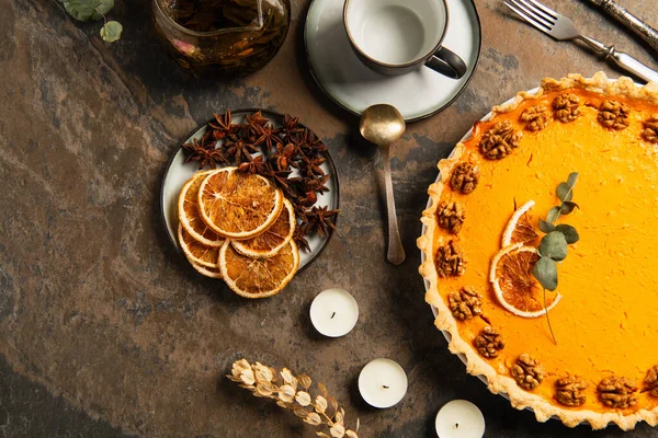 Tema de acción de gracias, pastel de calabaza adornado cerca de velas y rebanadas de naranja con especias en la mesa de piedra - foto de stock