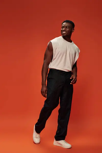 Hombre americano africano alegre y caliente en camiseta blanca y pantalones negros mirando hacia otro lado en el fondo rojo - foto de stock