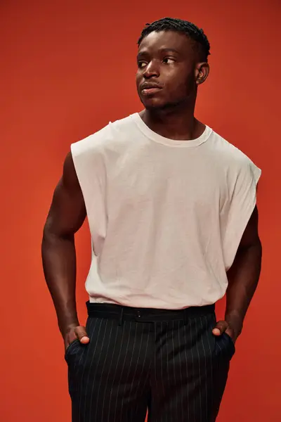 Modelo masculino afroamericano confiado y elegante cogido de la mano en bolsillos y mirando hacia otro lado en rojo - foto de stock