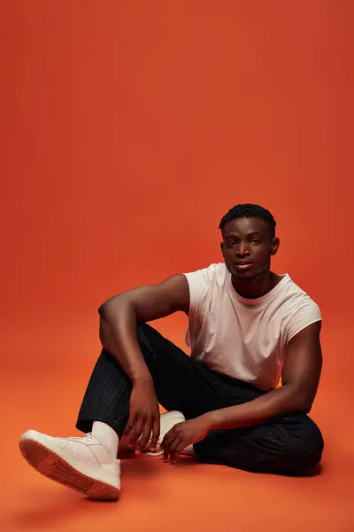 Afrikanisch-amerikanischer Mann in trendiger Freizeitkleidung sitzt und blickt in die Kamera auf rotem und orangefarbenem Hintergrund — Stockfoto