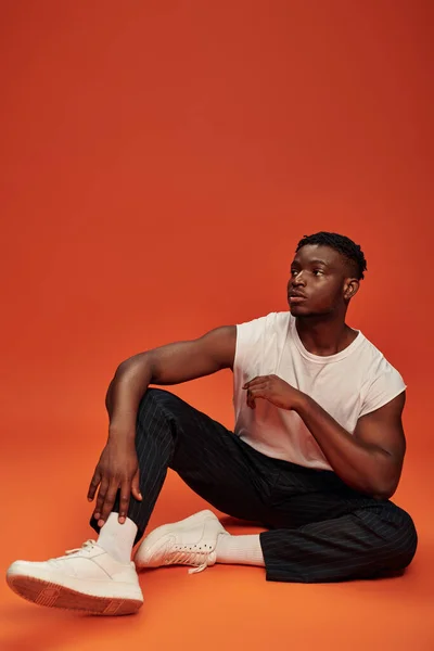 Full length of trendy african american guy sitting and looking away on red and orange backdrop — Stock Photo