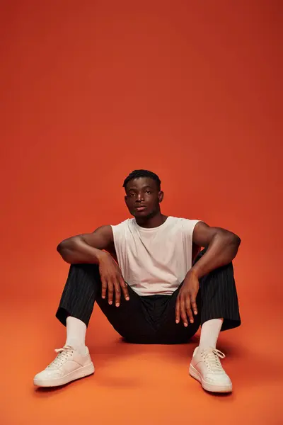 Confident and sportive african american man sitting and looking at camera on red and orange backdrop — Stock Photo