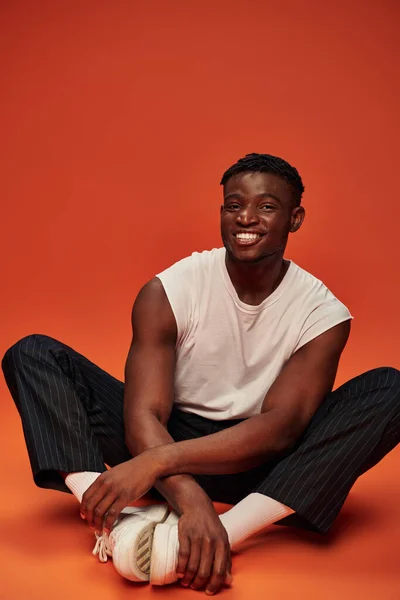 Young african american male model in white tank top and pants sitting an smiling at camera on red — Stock Photo