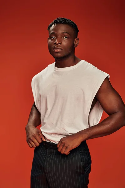 Hombre afroamericano pensativo en camiseta blanca y pantalones negros mirando hacia otro lado en el fondo rojo - foto de stock