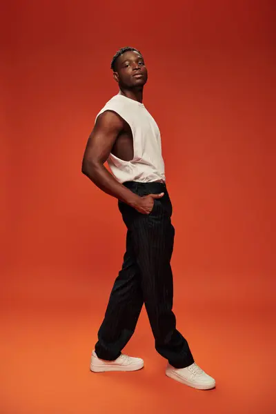 Hombre afroamericano seguro de sí mismo en camiseta blanca con la mano en el bolsillo de los pantalones negros en rojo - foto de stock