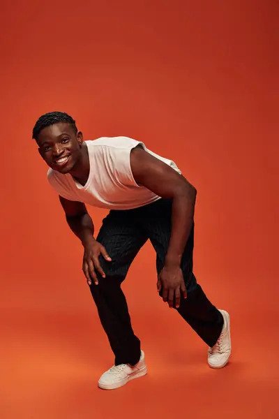 Happy african american guy in white tank top and pants leaning forward and looking at camera on red — Stock Photo