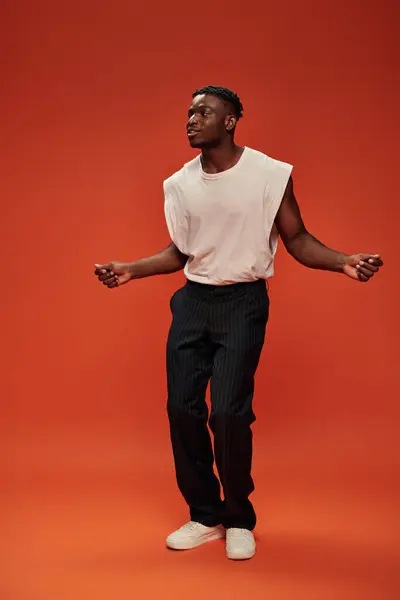 Asombrado y sonriente hombre afroamericano en ropa de moda mirando hacia otro lado en el fondo rojo y naranja - foto de stock