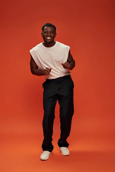 Hombre afroamericano lleno de alegría en ropa casual de moda posando sobre fondo rojo y naranja - foto de stock