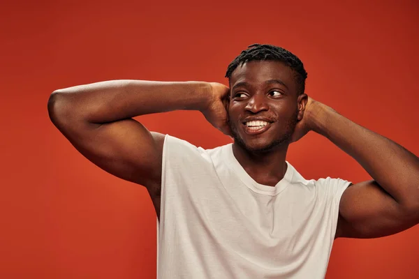 Athletic and joyous african american man in white tank top posing with hands behind head on red — Stock Photo