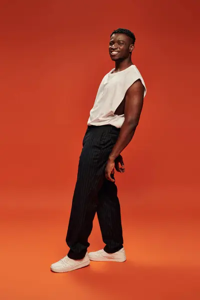 Despreocupado hombre afroamericano en pantalones negros y camiseta blanca posando sobre fondo rojo y naranja - foto de stock