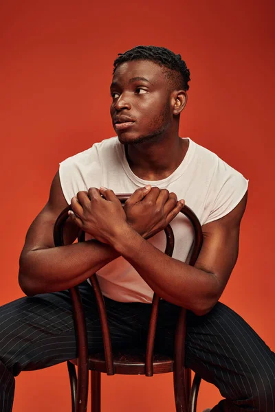 Young thoughtful african american man in white tank top sitting on chair and looking away on red — Stock Photo