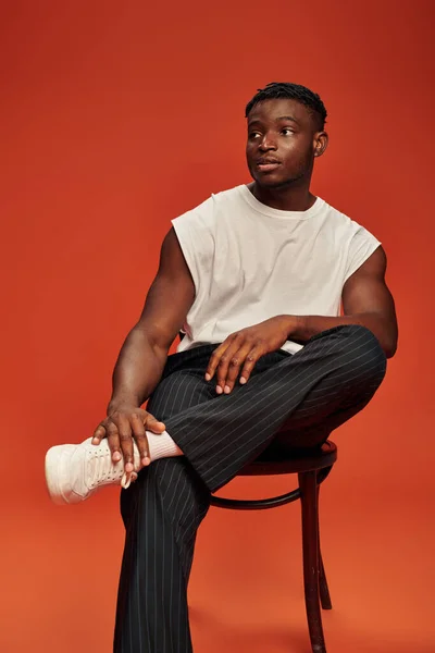 Chico afroamericano caliente en pantalones negros y camiseta blanca posando en la silla y mirando hacia otro lado en rojo - foto de stock
