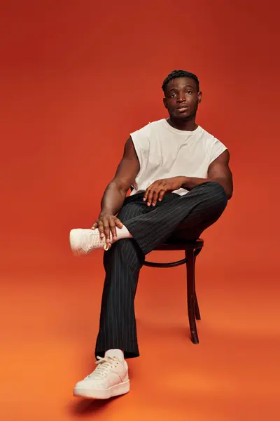 Афроамериканська чоловіча модель в стильному вуличному одязі, що сидить на стільці на червоному та помаранчевому тлі — стокове фото
