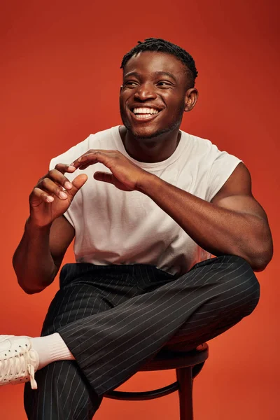 Laughing african american man in white tank top sitting on chair and gesturing on red backdrop — Stock Photo