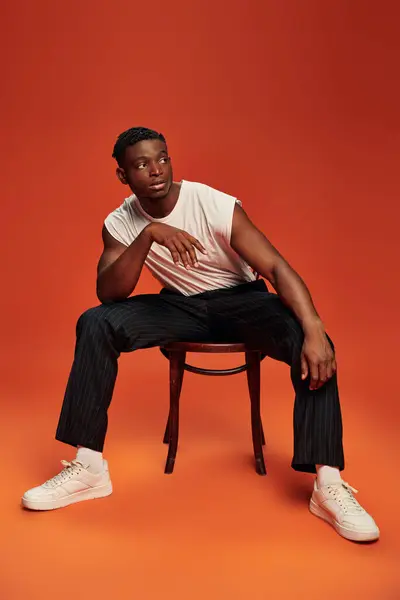 Сучасний і стильний афроамериканський чоловік сидить на стільці і дивиться на червоний і помаранчевий фон — стокове фото