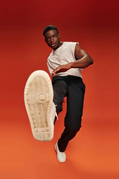 Hombre afroamericano con estilo en pose atrevida con la pierna extendida sobre fondo rojo y naranja - foto de stock