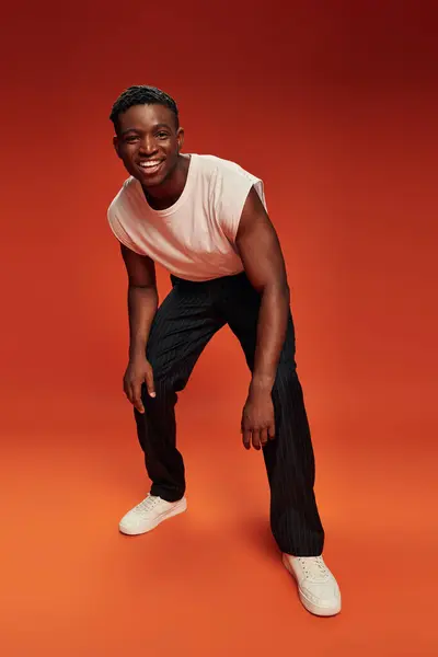 Longitud completa de hombre afroamericano de moda y feliz riéndose de la cámara en el fondo rojo y naranja - foto de stock