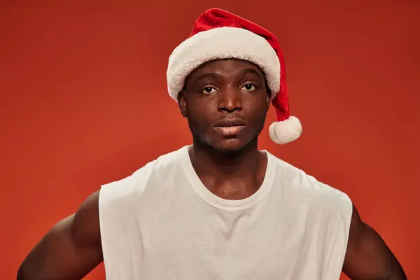 Hombre afroamericano serio en camiseta blanca y sombrero de santa mirando a la cámara en el fondo rojo - foto de stock