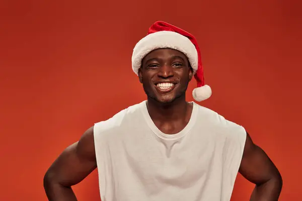 Cheerful african american man with radiant smile posing in santa hat and looking at camera on red — Stock Photo