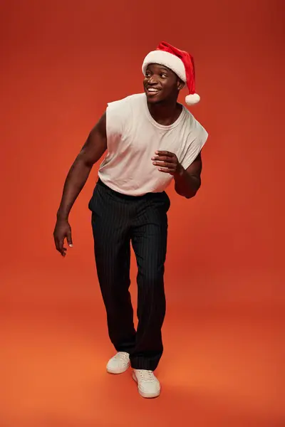 Cheerful african american guy in santa hat and stylish casual clothes looking away on red — Stock Photo