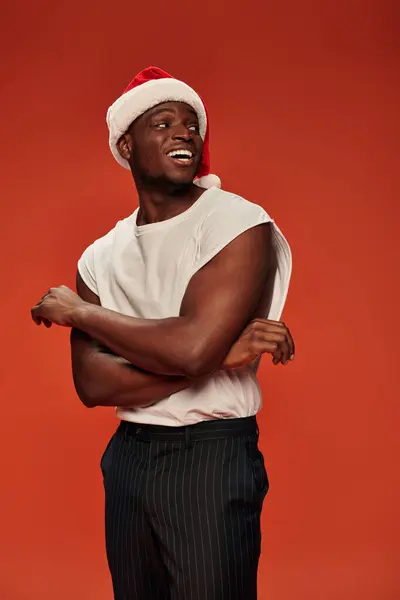 Hombre afroamericano emocionado en camiseta blanca y gorra de Navidad mirando hacia otro lado en el fondo rojo - foto de stock