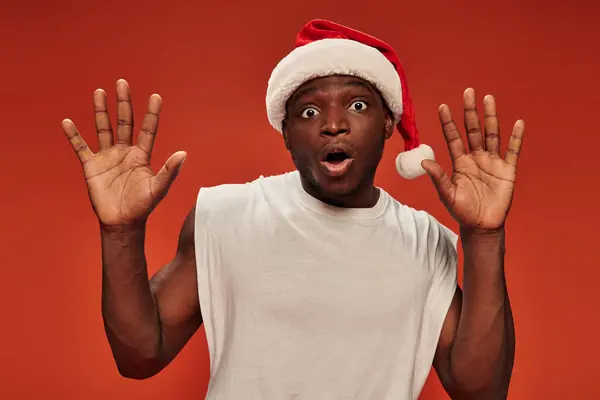 Scared african american guy in christmas hat and with open mouth showing stop gesture on red — Stock Photo