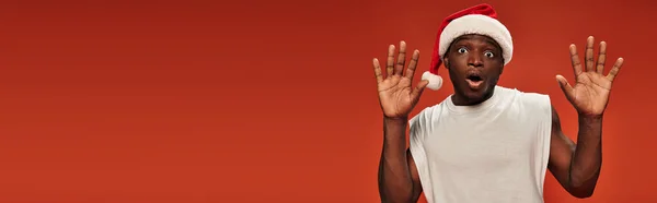 Frightened african american man in santa hat showing stop gesture with open mouth on red, banner — Stock Photo