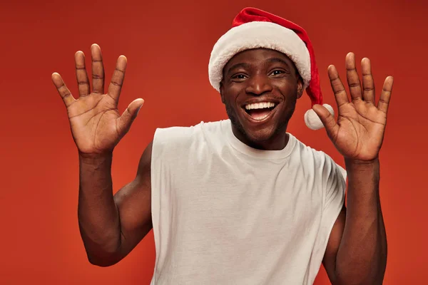 Joyful african american man in santa hat showing stop gesture with open mouth on red backdrop — Stock Photo