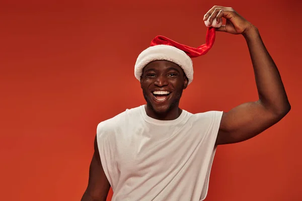 Joven y alegre hombre afroamericano tocando el sombrero de santa y sonriendo a la cámara en el fondo rojo - foto de stock