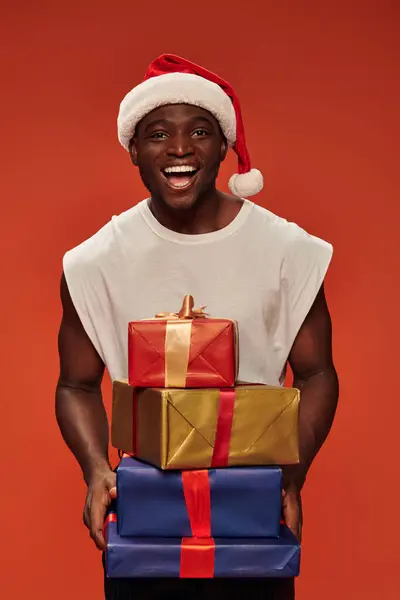 Allegro uomo afro americano in cappello di Babbo Natale con scatole regalo colorate sul rosso, auguri di vacanza — Foto stock