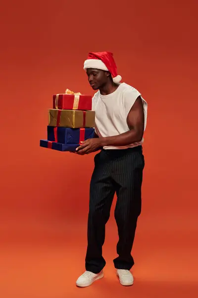 Heureux homme afro-américain en chapeau de Père Noël festif tenant des cadeaux de Noël colorés sur rouge — Photo de stock
