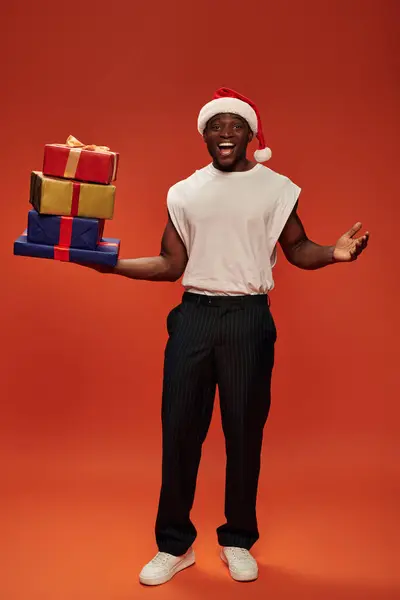 Emocionado afroamericano chico en santa gorra celebración de cajas de regalo y mostrando wow gesto sobre fondo rojo - foto de stock