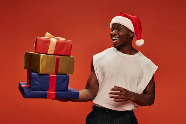 Amazed and overjoyed african american man in santa hat looking at colorful gift boxes on red — Stock Photo