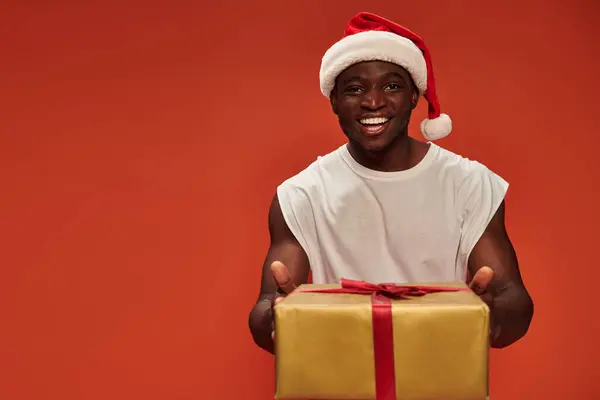 Задоволений афроамериканський чоловік у різдвяному капелюсі, що показує подарункову коробку зі святковим подарунком на червоному — стокове фото