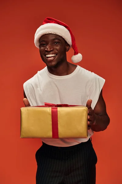 Cheerful african american guy in christmas hat holding holiday gift and smiling at camera on red — Stock Photo