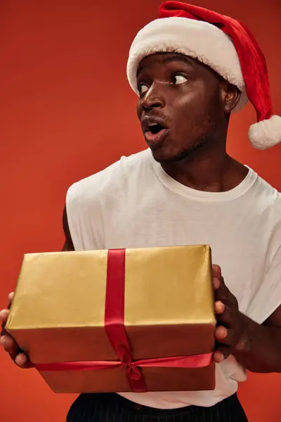 Amazed african american man in santa hat holding gift box and looking away with open mouth on red — Stock Photo